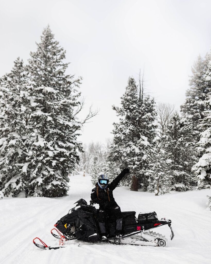 Person on snowmobile waving in snowy forest scene.