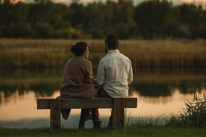 Couple sitting on a wooden bench by a calm river at sunset in fall